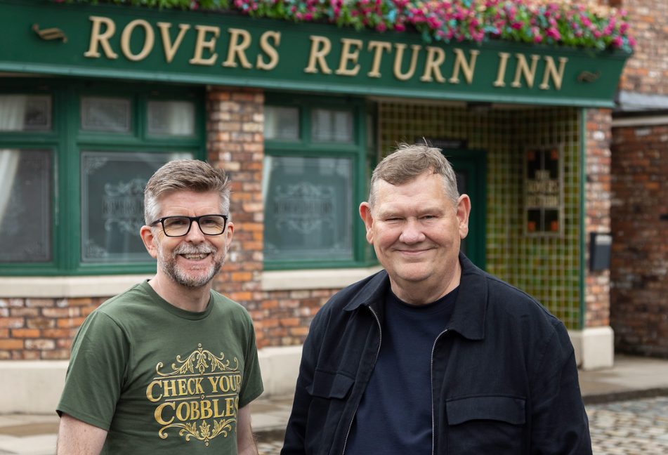 Two people outside the Rovers Return Inn on the Coronation Street set supporting Macmillan Cancer Support's Check Your Cobbles campaign.