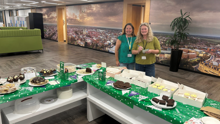 Two Aviva employees standing behind a large spread set out for a Coffee Morning at their offices.