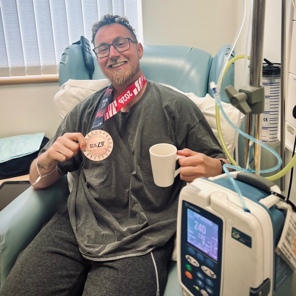 Gary is sitting down in a hospital chair receiving treatment. He is holding in his hand a finisher's medal for the London marathon. In his other hand is a coffee mug.