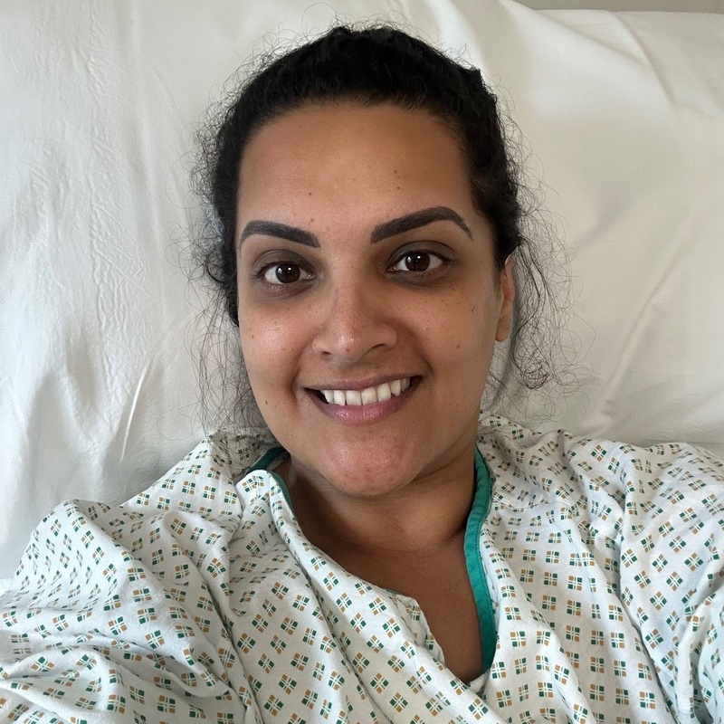 A person sitting in a hospital bed wearing a hospital gown and looking towards the camera during cancer treatment.
