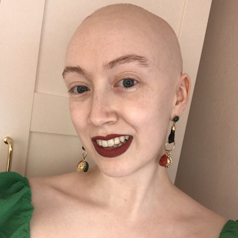 Portrait of a person with a shaved head wearing a green top and statement earrings, smiling for a close-up photograph.