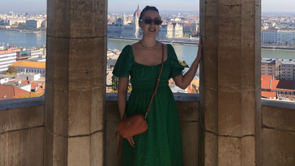 A person in a green dress stands beneath stone arches overlooking a riverside city skyline, with historic buildings and a clear blue sky in the background.