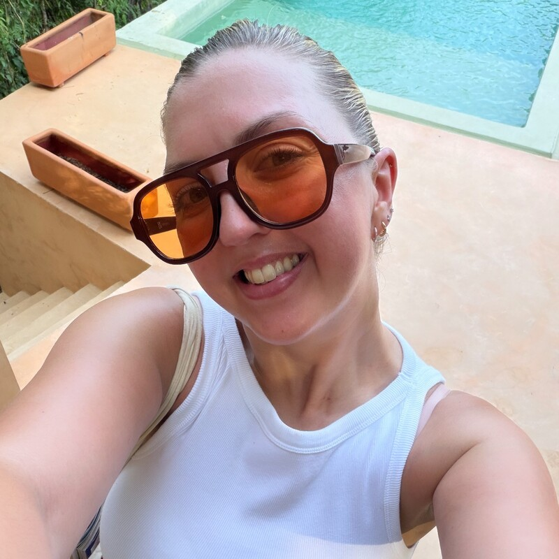 A selfie of a person wearing oversized sunglasses, a white sleeveless top and red trousers, standing beside an outdoor pool surrounded by greenery.