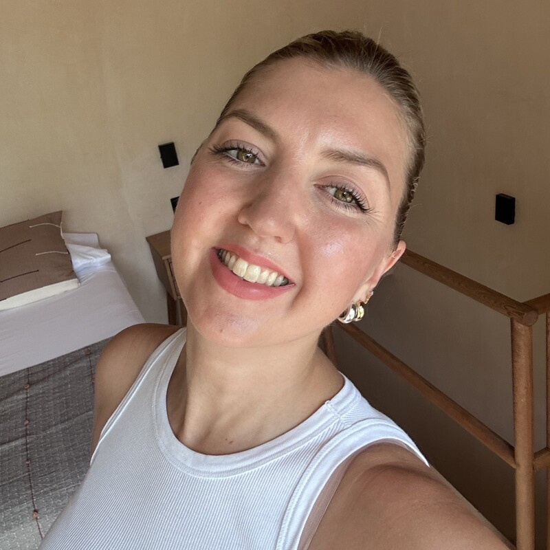 A selfie of a person wearing a white sleeveless top in a bright hotel room, with a bed and minimalist décor visible in the background.