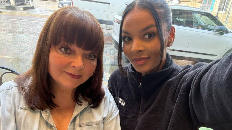 A person takes a selfie with another person while sitting indoors beside a large window. A street scene with parked vehicles is visible outside in the background. The image captures a moment together in a casual setting.