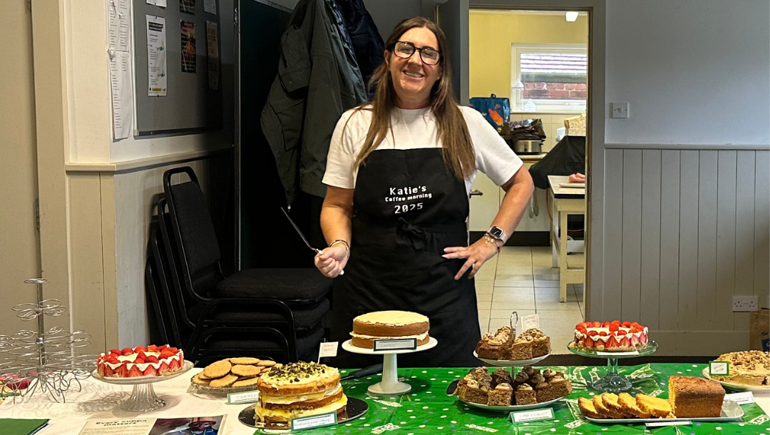 Katie is standing in front of a table prepared for a Coffee Morning. In one hand she is holding a pie server utensil. The table is full of cakes beautifully decorated. Katie is wearing a black apron and one T-shirt.