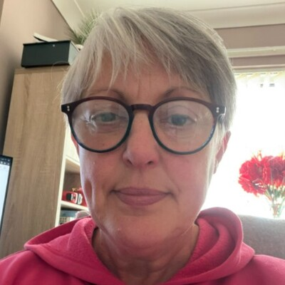 A close-up portrait of a person with short grey hair and round glasses, wearing a bright pink hoodie and sitting indoors in a home office setting with natural light and red flowers visible in the background.
