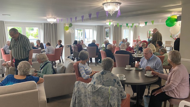 A Coffee Morning at a Churchill lodge. Residents are gathered around a large lounge, some are sitting down on comfy chair. Others are sat around tables enjoying warm drinks like tea and coffee. There is colourful bunting and balloons decorating the room.