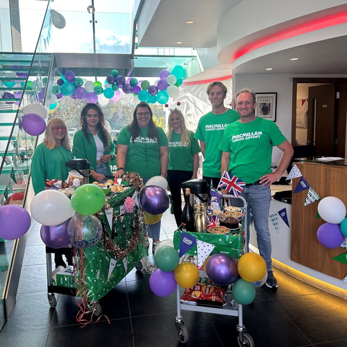 A group of people celebrating Coffee Morning. They are standing side by side wearing green tops. In front of them are two trolleys decorated with Coffee Morning, and includes yummy food and drinks.
