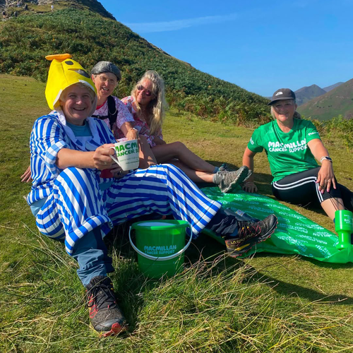 Becky is dressed as Bananas in Pyjamas at the top of Cowbell in the Lake District. She is sat down on a grassy hill with others.