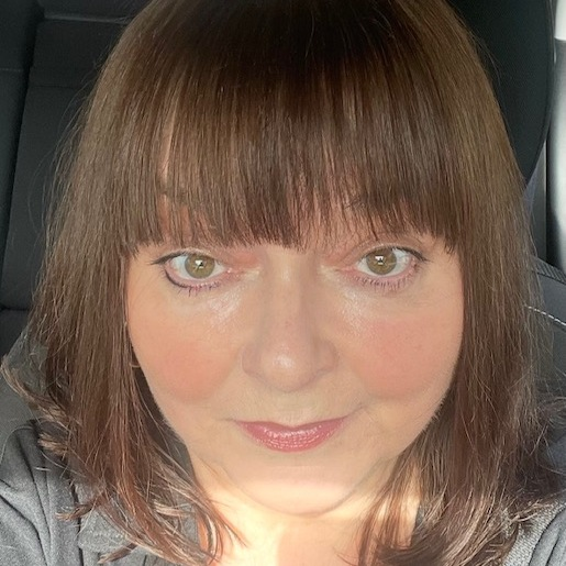 Close-up portrait of a person with shoulder-length dark brown hair and a fringe, looking directly at the camera while seated in a car. Natural daylight highlights the face, creating a warm and candid image.