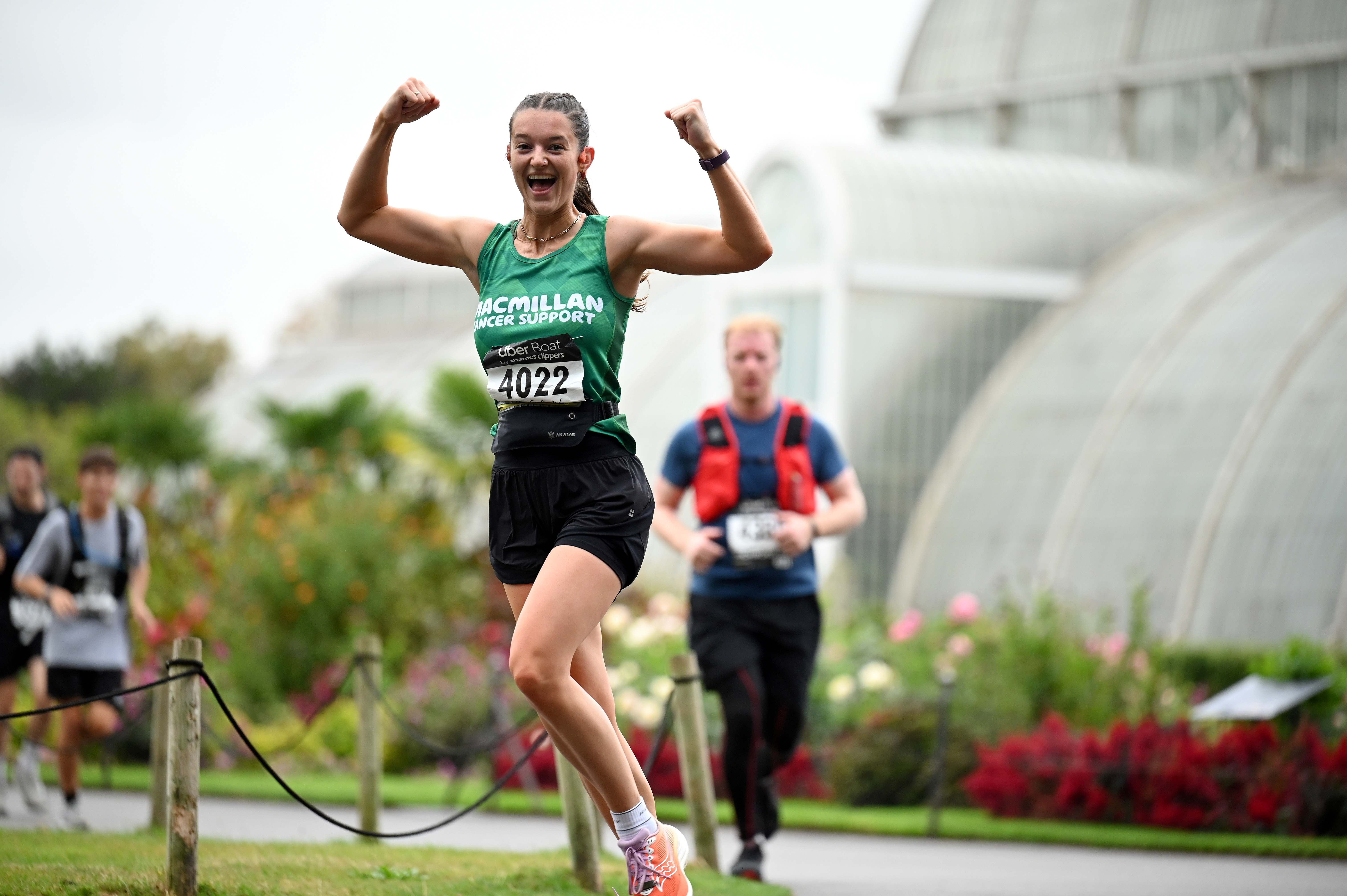 A woman wearing a Macmillan running vest running and flexing her muscles as she runs past a glass house in Kew Gardens.