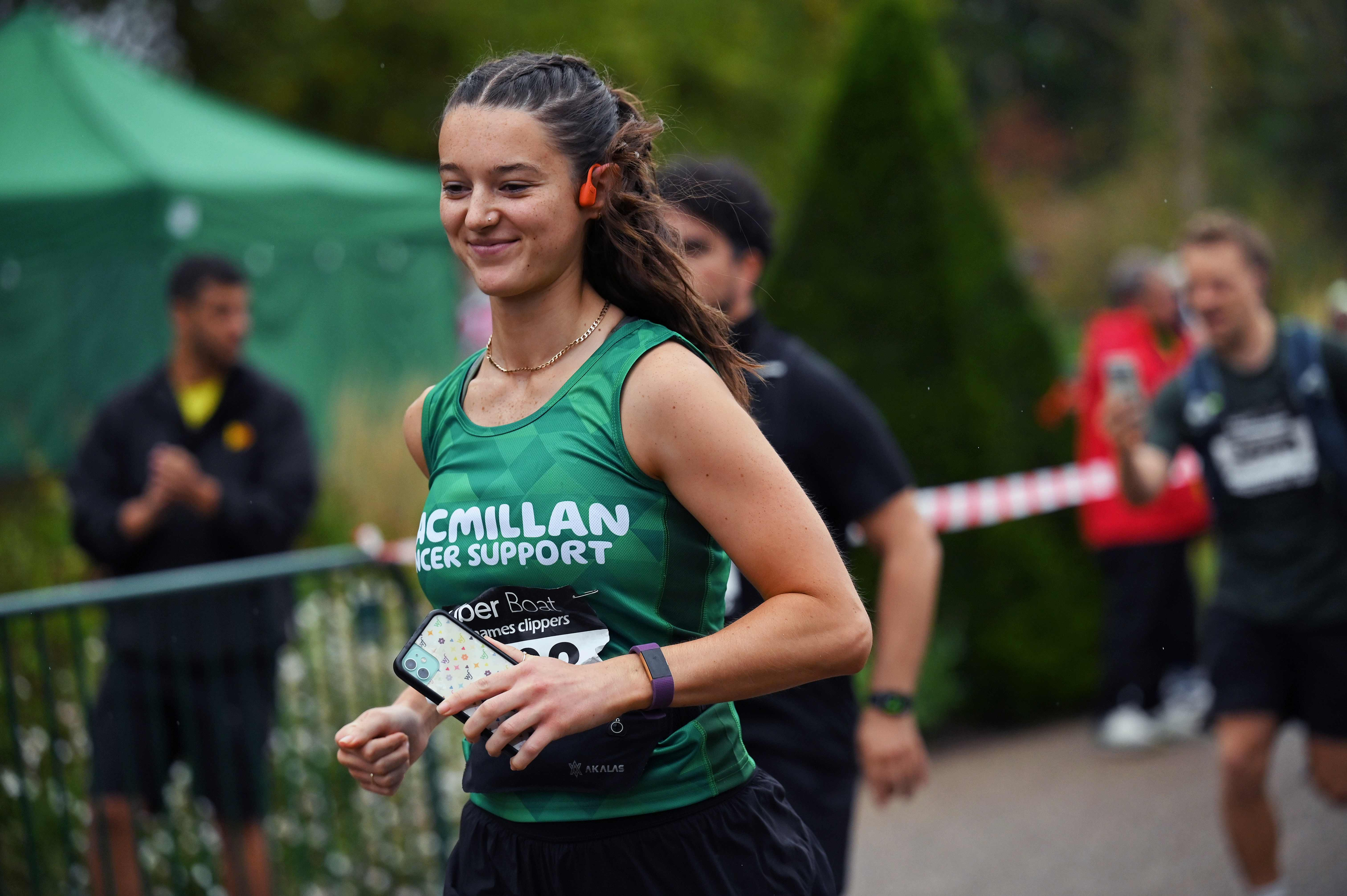 A woman wearing earphones and smiling as she runs in a Macmillan running vest and holding her phone.