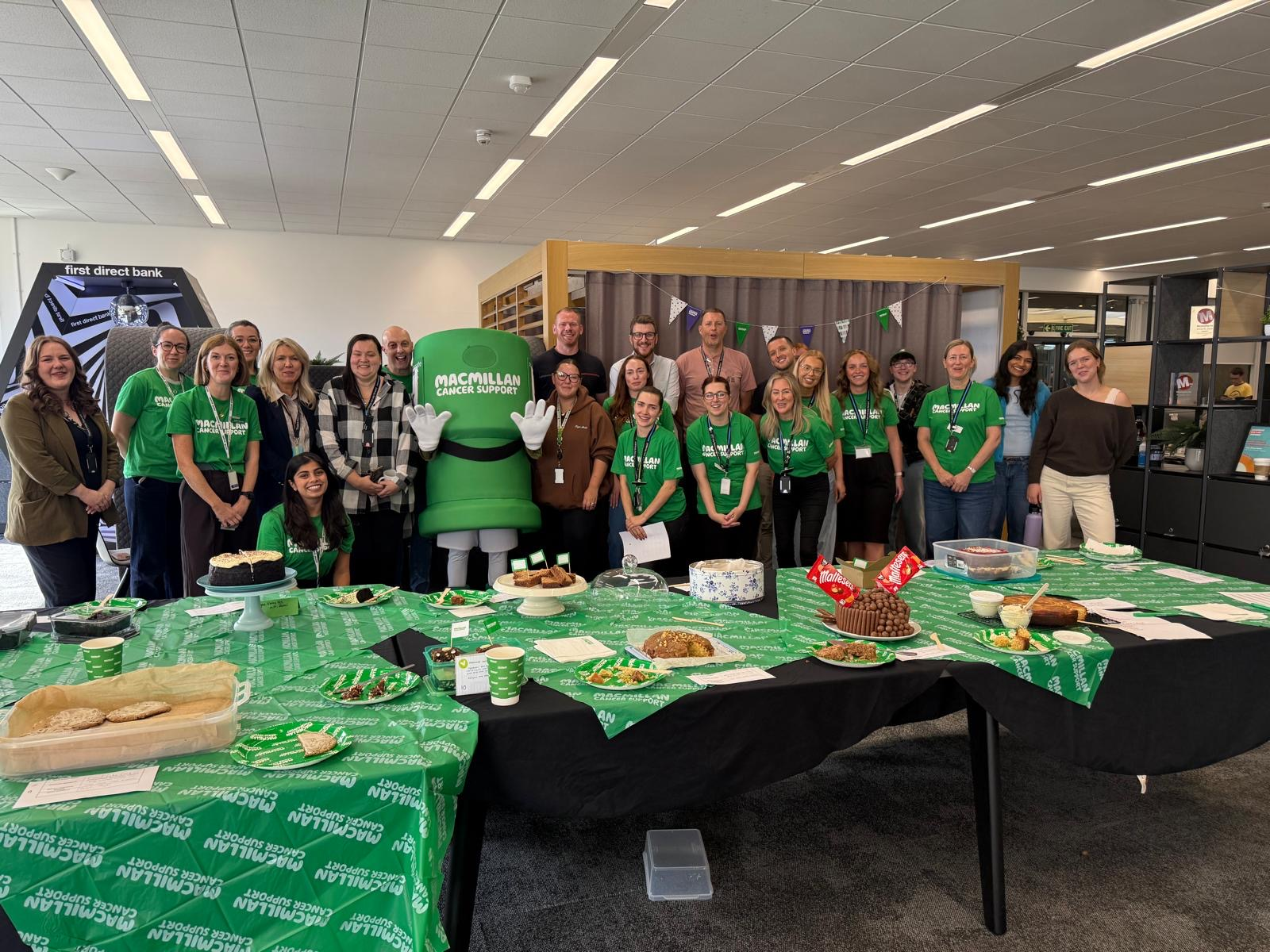 A group of people gathered around a Macmillan Cancer Support mascot and a table of cakes and refreshments at a fundraising event.