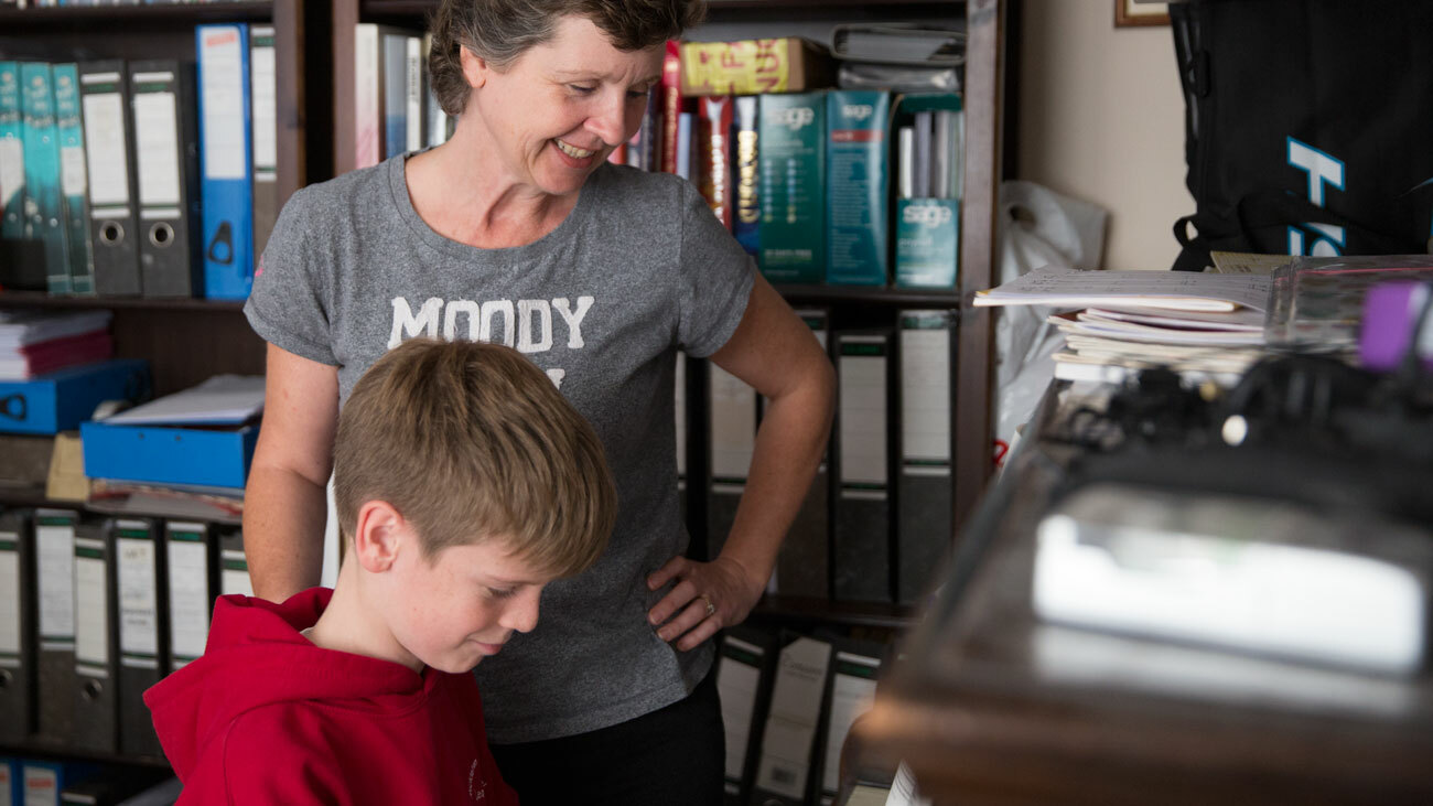Diane is stood watching her son on the piano in what appears to be her home study. She is wearing a grey tshirt and is smiling. 