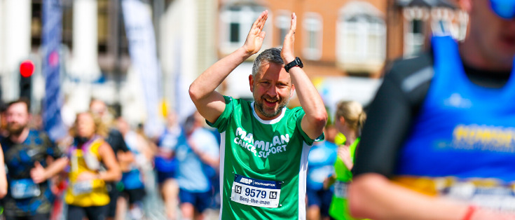 Man applauding with his hands above his head as he runs while wearing a green Macmillan t-shirt.