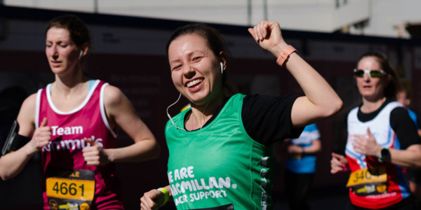 Woman running in a green Macmillan running vest over a black t-shirt punching the air and smiling.