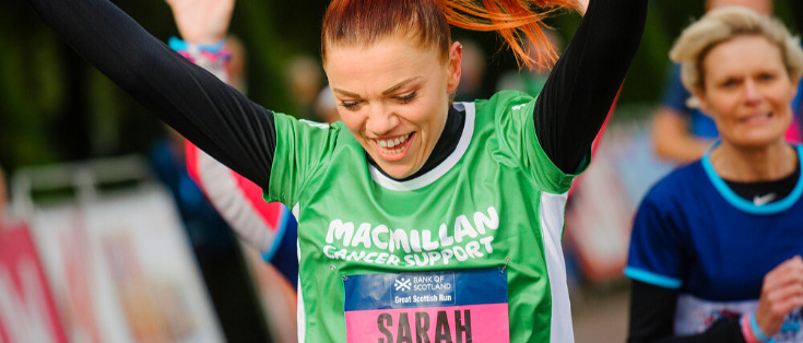 A woman with Sarah written on her green Macmillan running top smiling with her hands in the air.