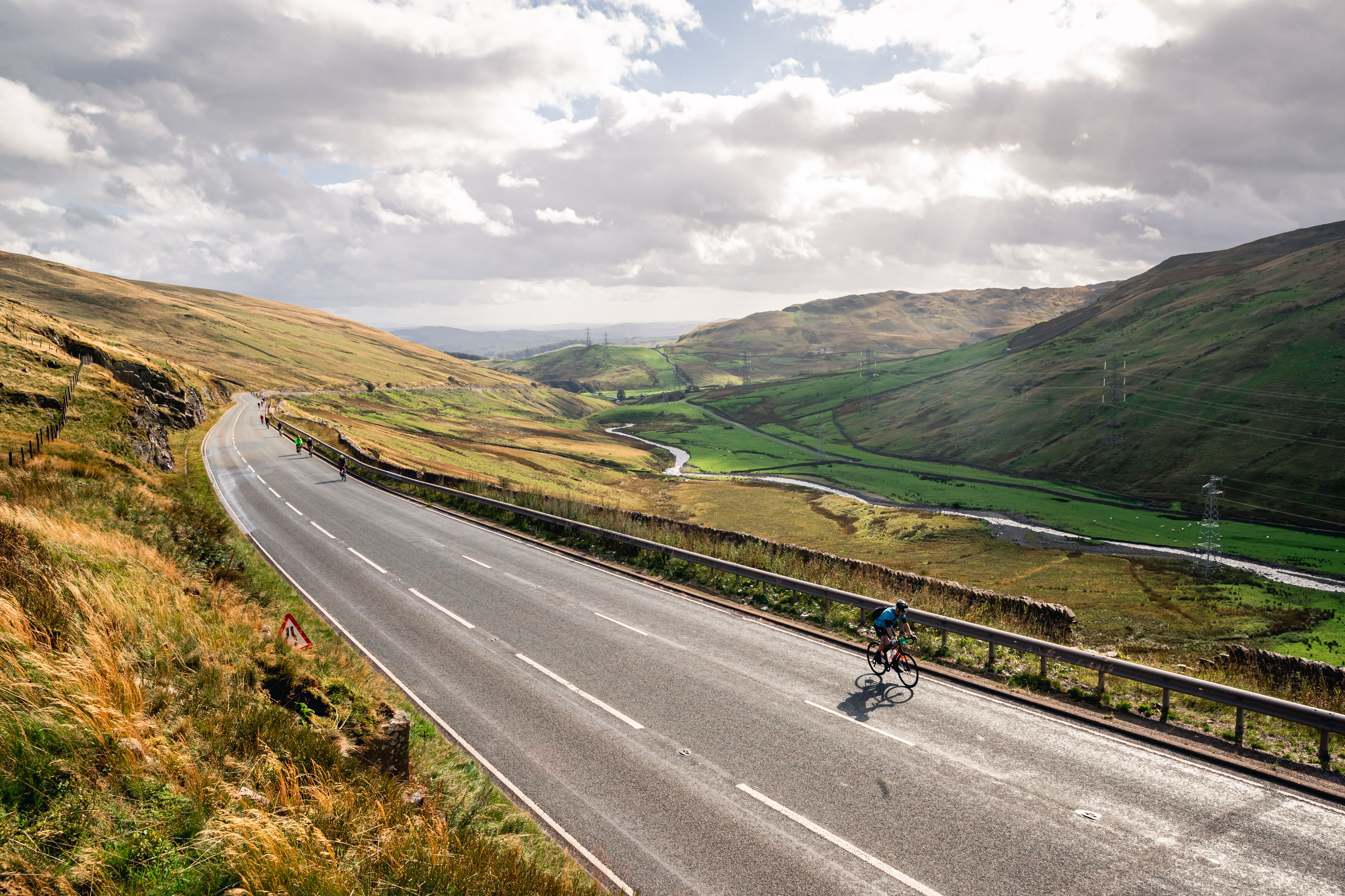 Someone cycling up an empty winding road. There is a river running through a valley and large hills in the background.
