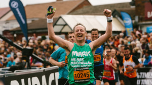 Mark, wearing a green Macmillan running vest with both arms in the air in celebration at the end of a race.