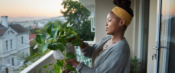 A landscape profile of a Black woman on a balcony overlooking a residential street. She is holding a potted plant in her hand. 
