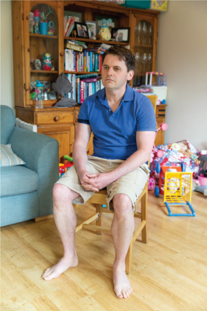 Position 1. A man sits upright on a chair. His arms and shoulders are relaxed. His feet are flat on the floor.