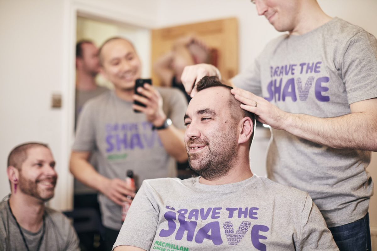 A man's head is being shaved by another man for charity. They are wearing 'Brave the Shave' teachers.