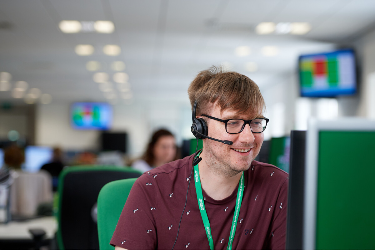 A Macmillan Support Line advisor. The man with blonde hair is smiling and wearing a headset and a patterned top.