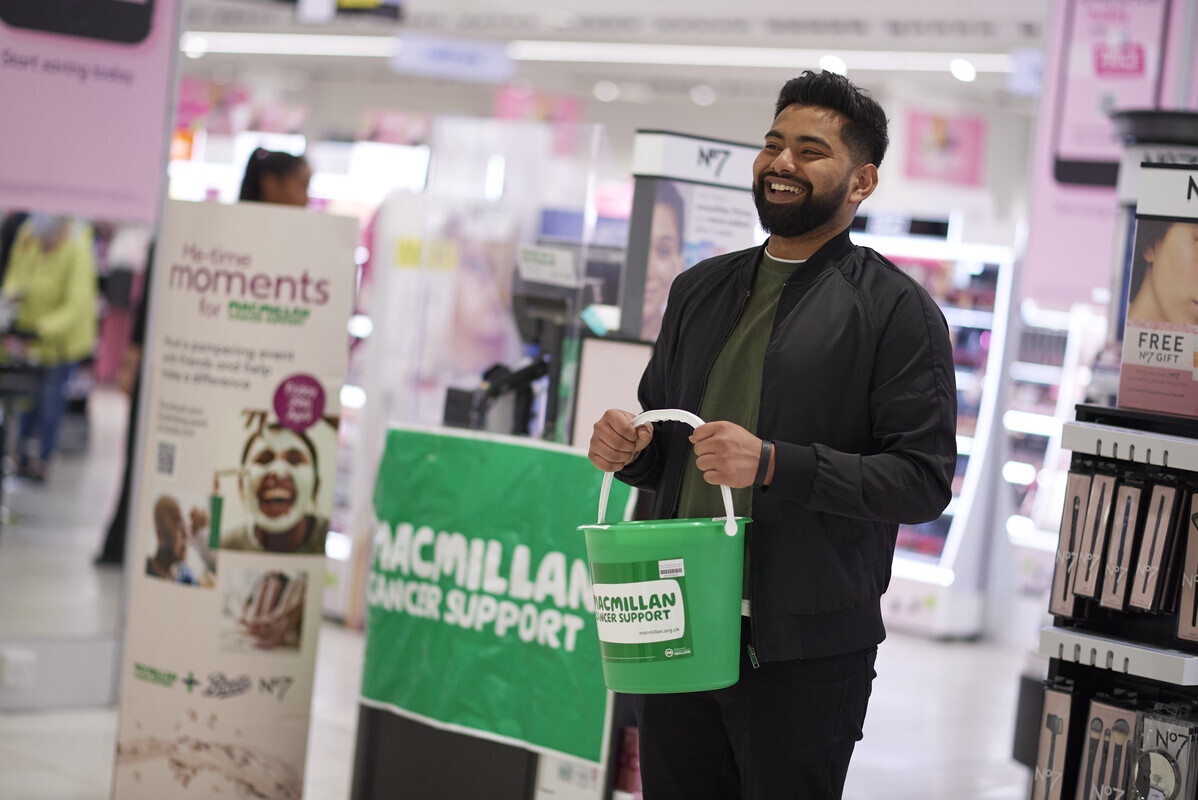 A Macmillan volunteer holds a green Macmillan fundraising bucket in a shop