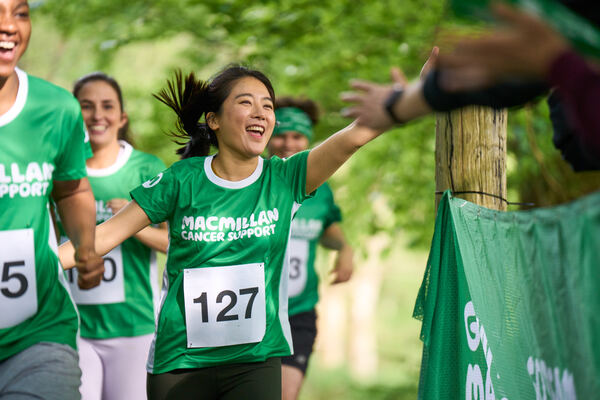 A woman in a green Macmillan running tshirt runs alongside other women, smiling and waving at supporters