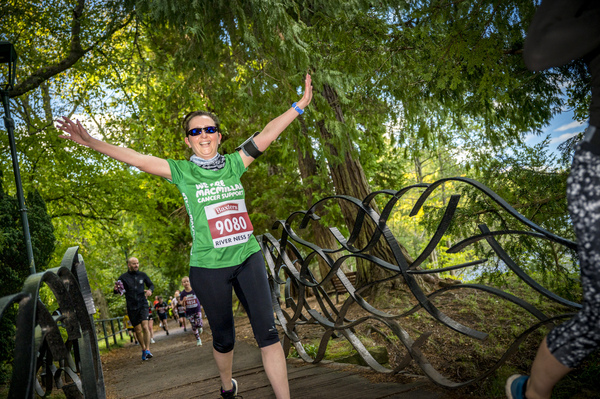 A woman jogging across a small bridge with her arms in the air, wearing a green Macmillan running t-shirt