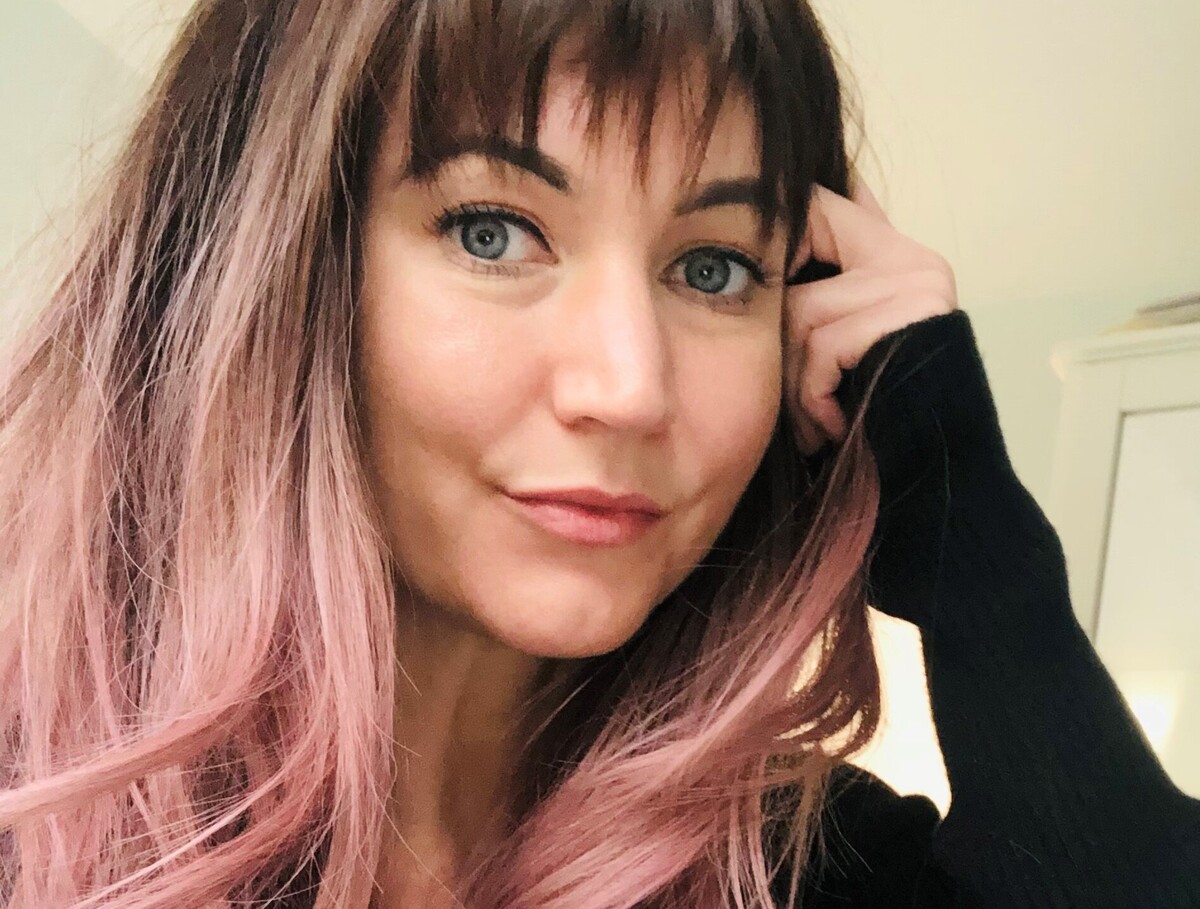 A lady with long pink hair wearing a black jumper and wearing jeans.