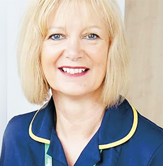 Headshot of a blonde white woman smiling. She is wearing a blue nurse uniform '